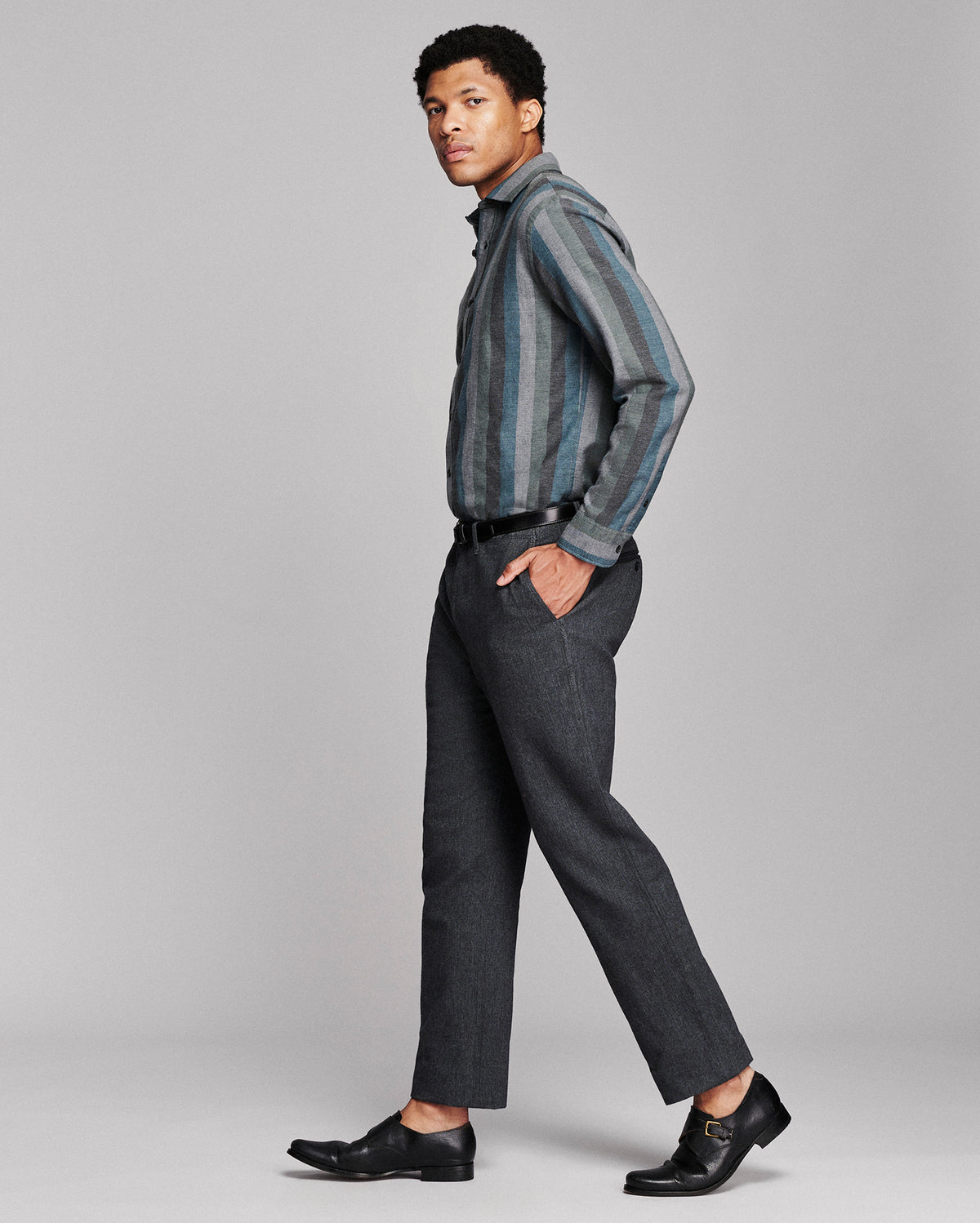 A man modeling a charcoal gray striped button-down shirt and dark gray trousers with black dress shoes, photographed against a solid light gray studio background.