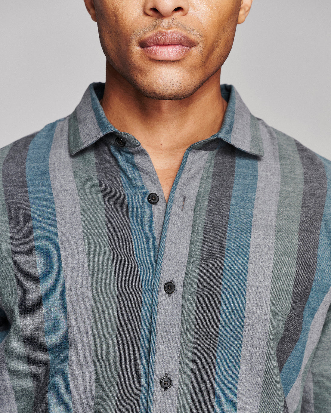Close-up of a man wearing a button-front collared shirt with vertical stripes in muted blue-gray, gray, and taupe tones against a plain light gray background.
