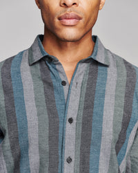 Close-up of a man wearing a button-front collared shirt with vertical stripes in muted blue-gray, gray, and taupe tones against a plain light gray background.