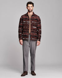 A man modeling a patterned button-up shirt jacket over a tan zip-up sweater, paired with gray trousers and brown suede shoes, against a solid gray studio background.