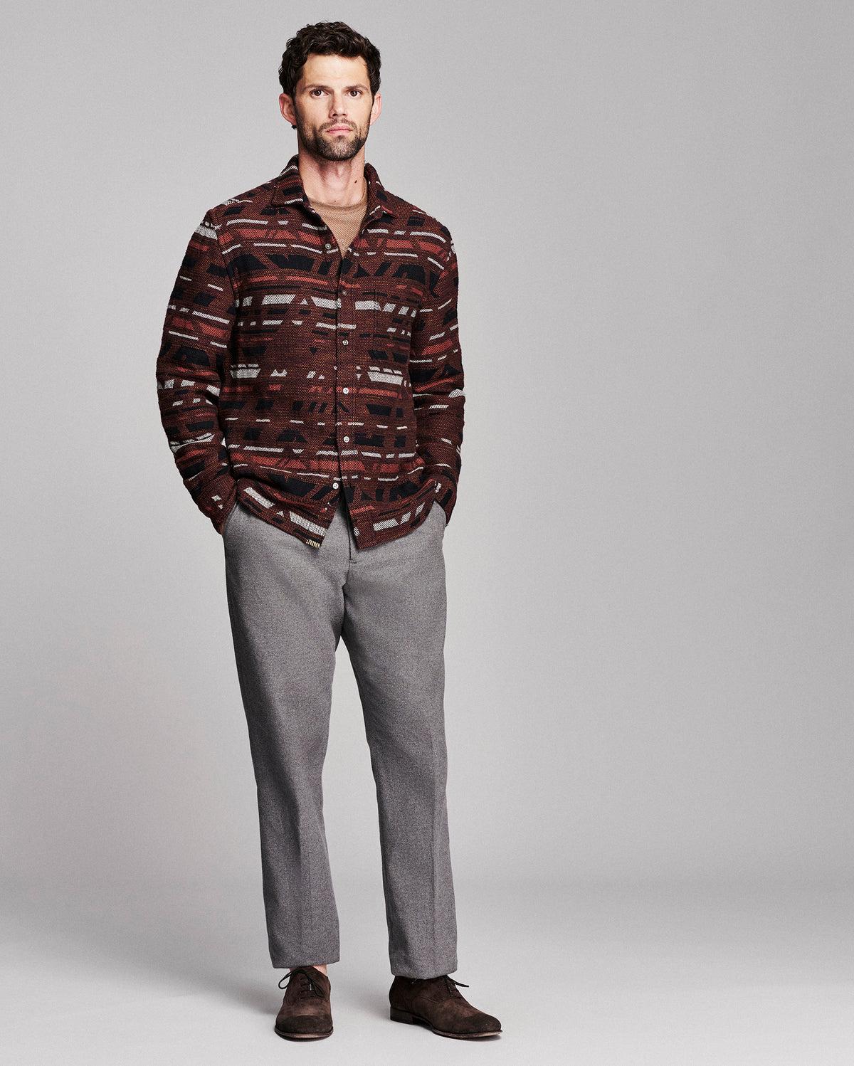 A man modeling a long-sleeve button-up shirt with a dark brown, red, and black abstract pattern, paired with gray trousers and brown shoes, against a solid gray studio background.