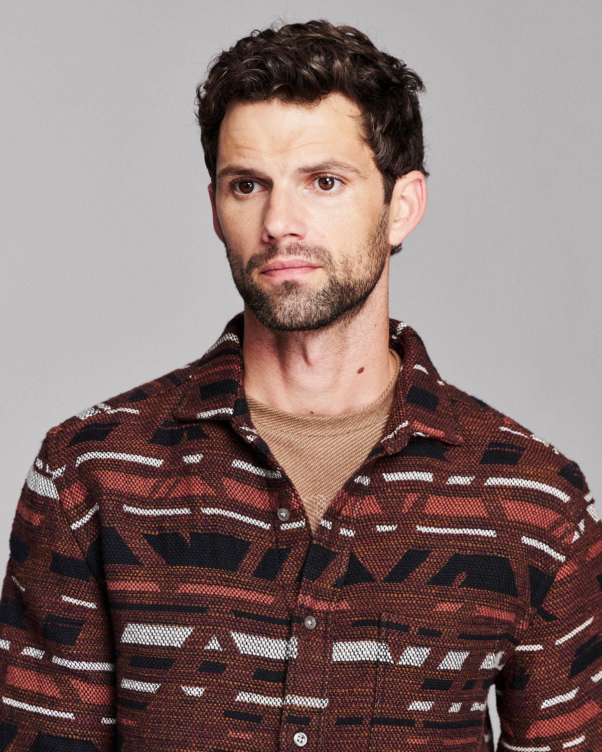 A man wearing a brown and black patterned button-down shirt over a beige undershirt, photographed against a solid gray background.