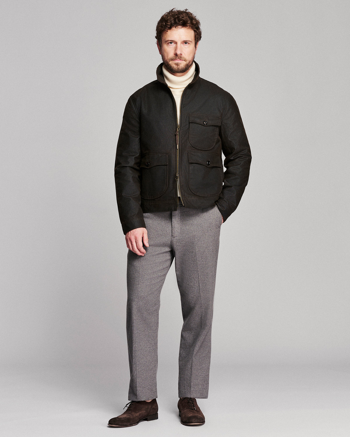 A man modeling a black zip-up jacket over a cream turtleneck sweater, paired with gray trousers and dark brown shoes, against a solid light gray studio background.