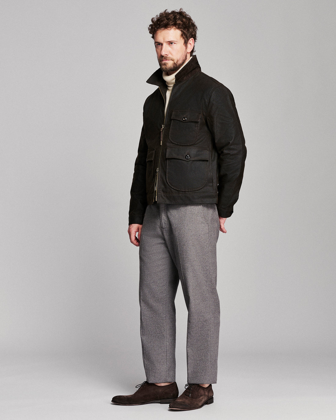 A man modeling a black utility jacket over a cream turtleneck, paired with gray wool trousers and dark brown leather shoes, shot against a solid light gray studio background.
