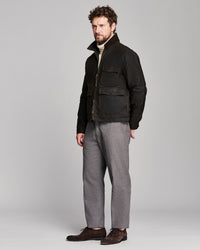 A man modeling a black utility jacket over a cream turtleneck, paired with gray wool trousers and dark brown leather shoes, shot against a solid light gray studio background.