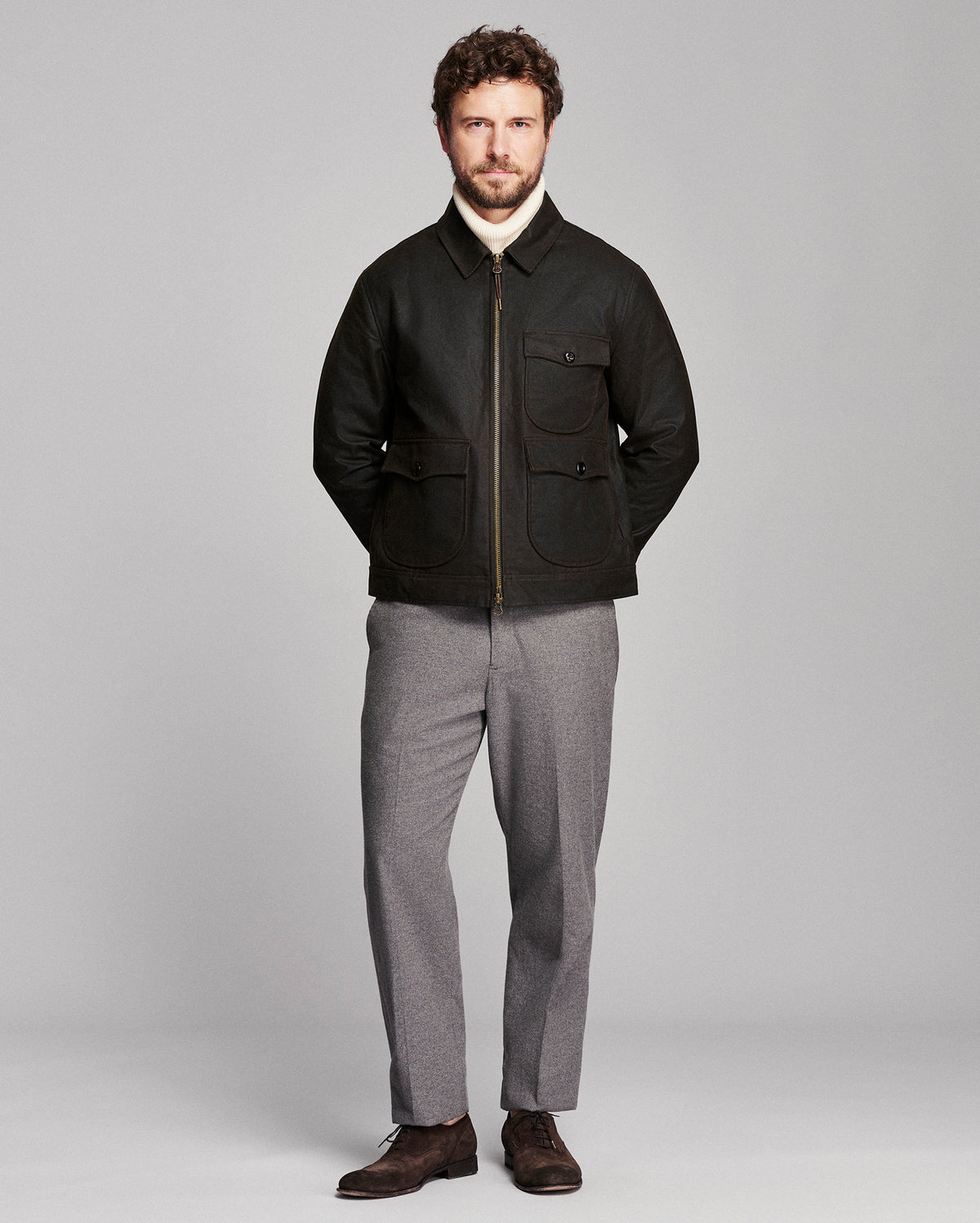 A man modeling a black zip-up jacket with front flap pockets, worn over a white turtleneck and paired with gray trousers and dark brown shoes, against a light gray studio background.