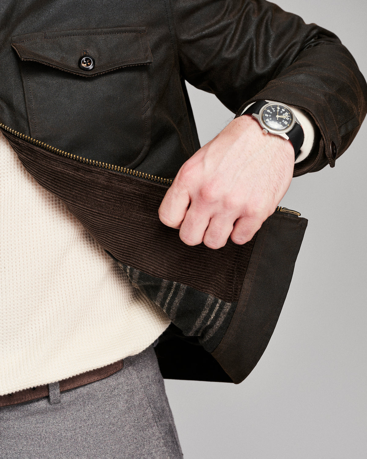 Close-up of a person wearing a dark brown leather jacket with a corduroy zippered panel, a cream knit sweater underneath, gray trousers, and a wristwatch.