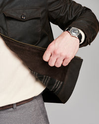 Close-up of a person wearing a dark brown leather jacket with a corduroy zippered panel, a cream knit sweater underneath, gray trousers, and a wristwatch.