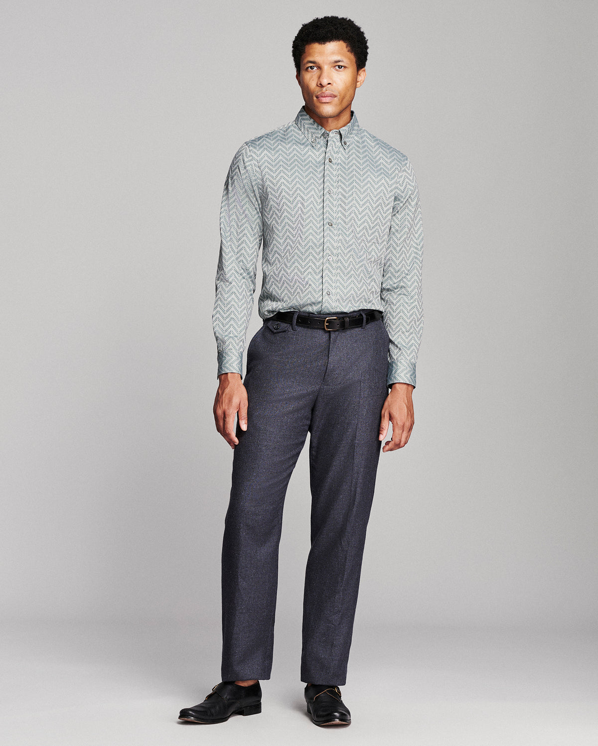 A man modeling a light gray and white chevron-patterned button-down shirt paired with dark gray trousers, a black belt, and black dress shoes, against a neutral gray studio background.