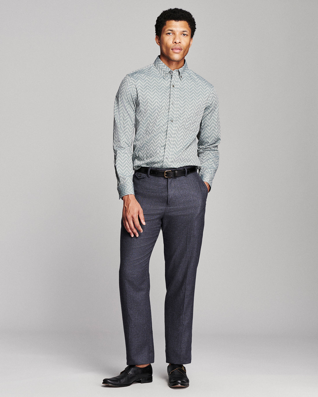 A man modeling a light gray and white chevron-patterned button-down shirt with dark gray trousers, a black belt, and black dress shoes, standing against a light gray studio background.