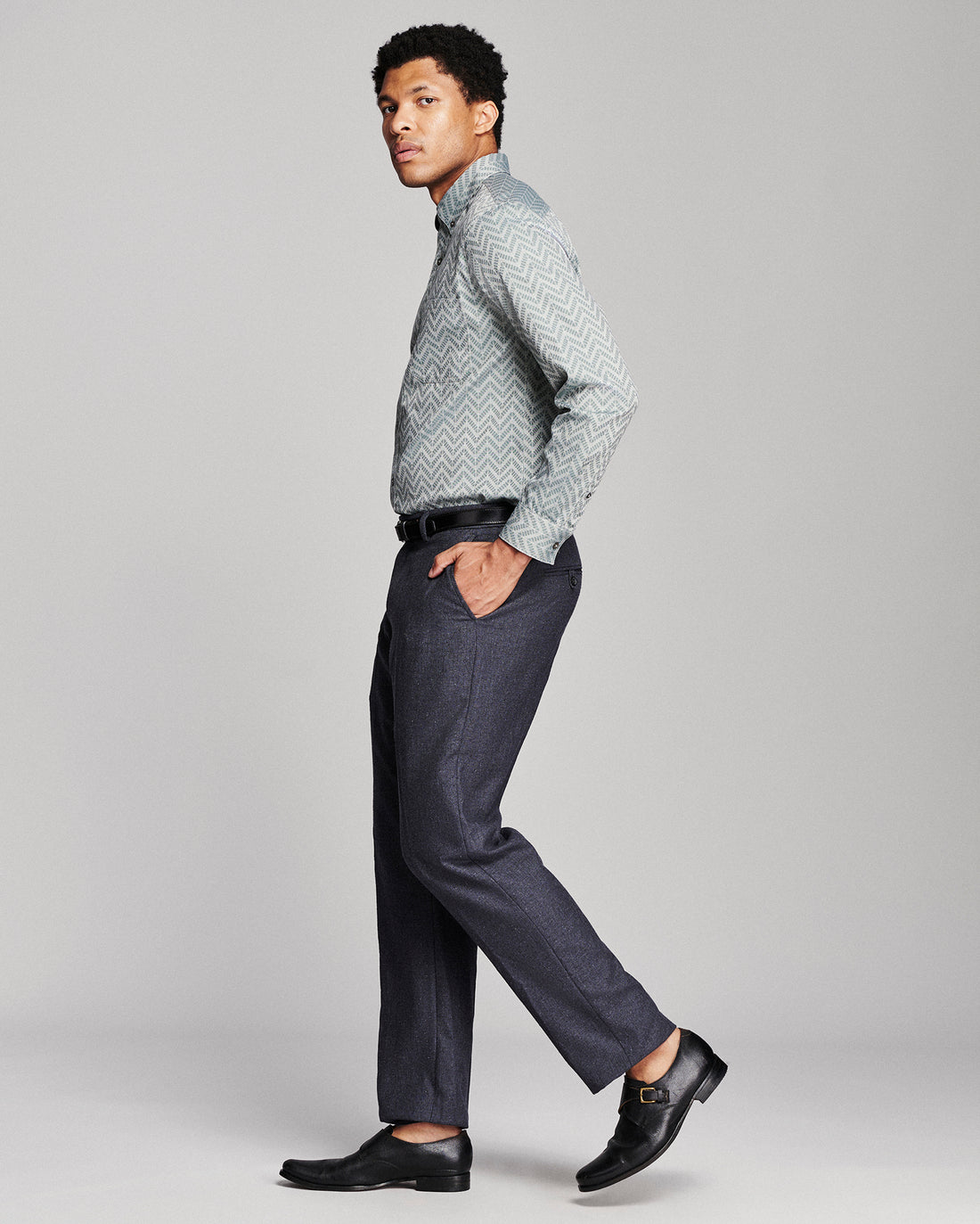 A man modeling a light gray and white patterned long-sleeve button-down shirt, paired with dark gray trousers and black leather dress shoes, shown in profile against a light gray studio background.
