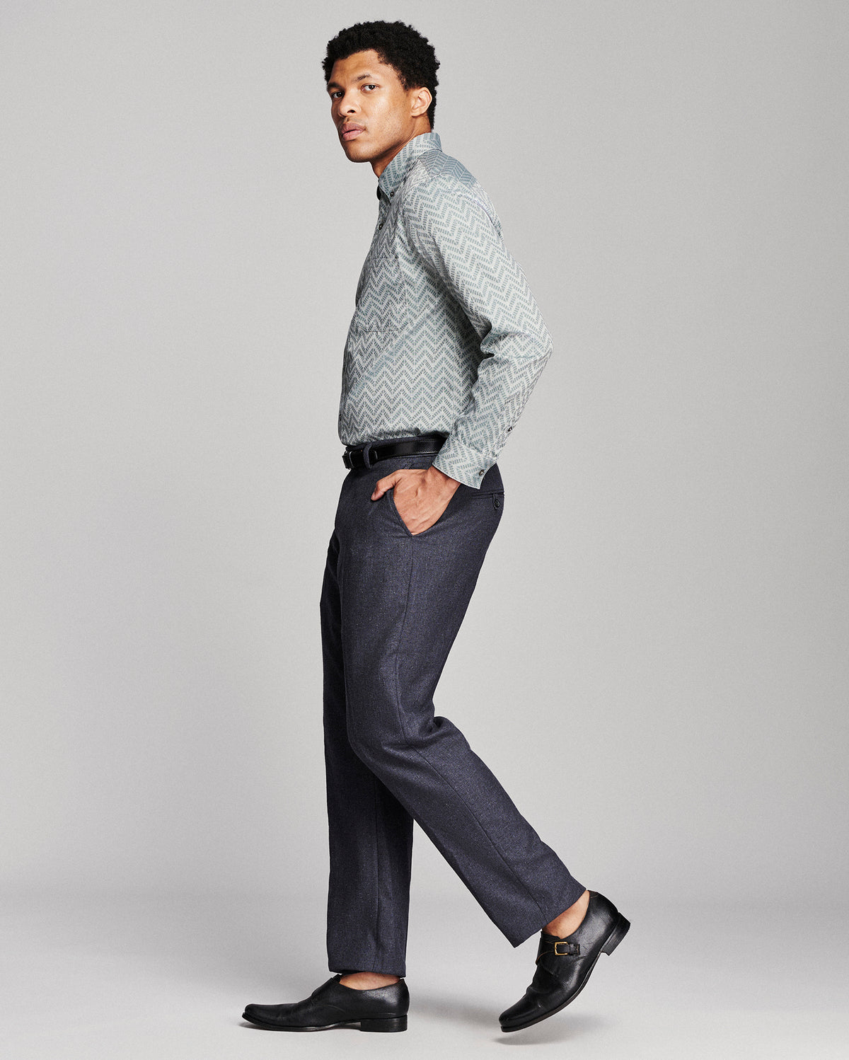 A man modeling a light gray and white patterned long-sleeve button-down shirt, paired with dark gray trousers and black leather dress shoes, shown in profile against a light gray studio background.