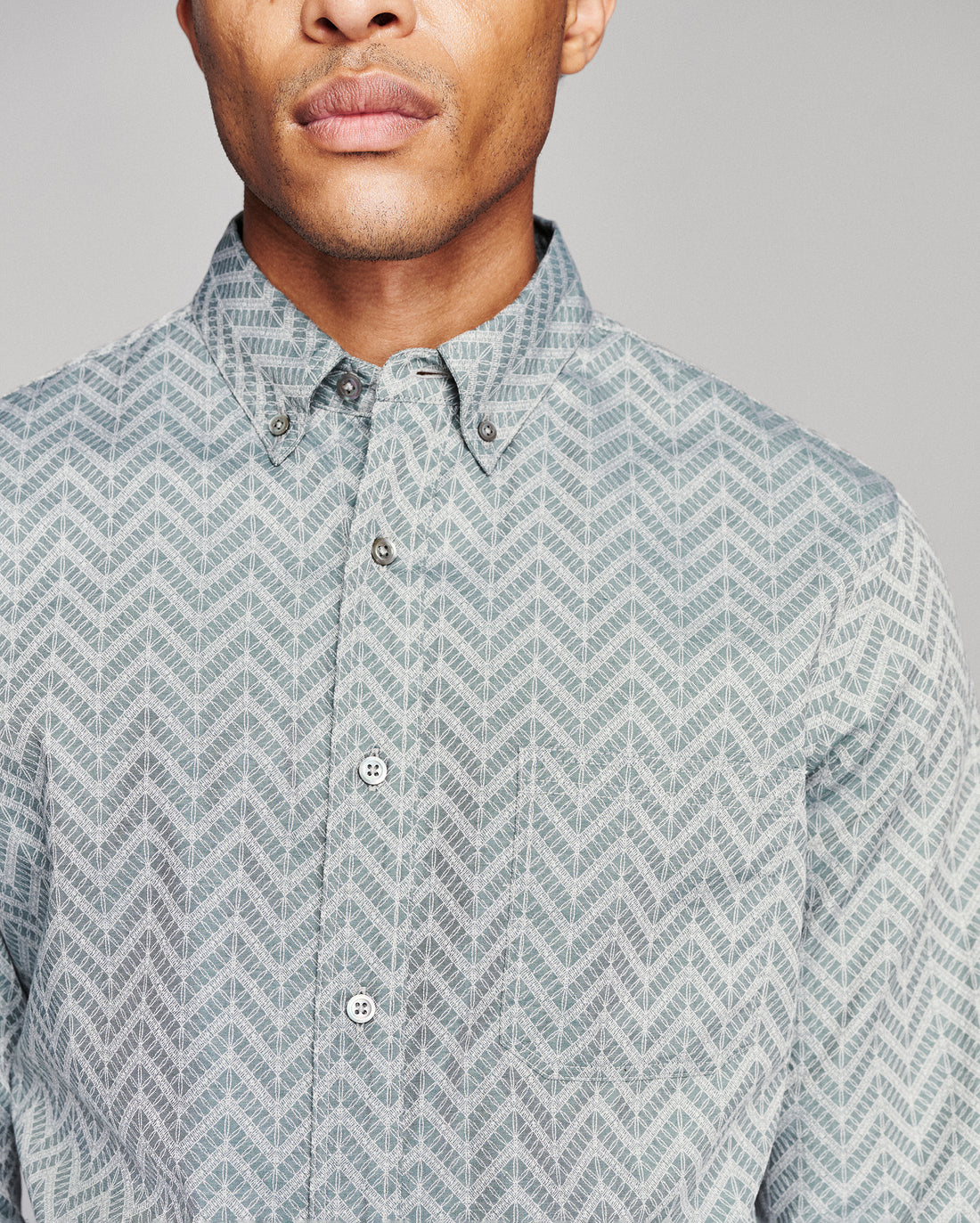 A close-up of a man wearing a light gray and white patterned button-down shirt, showing the collar, front buttons, and part of the chest against a plain gray background.
