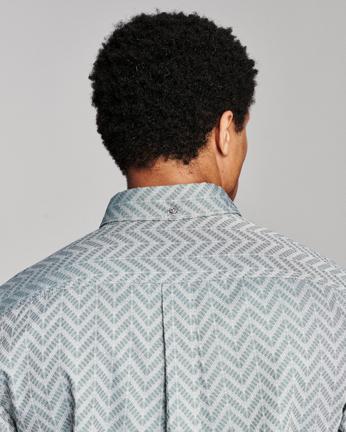 Back view of a man wearing a light blue and gray patterned button-down shirt, showing the collar and shoulder area against a solid gray background.