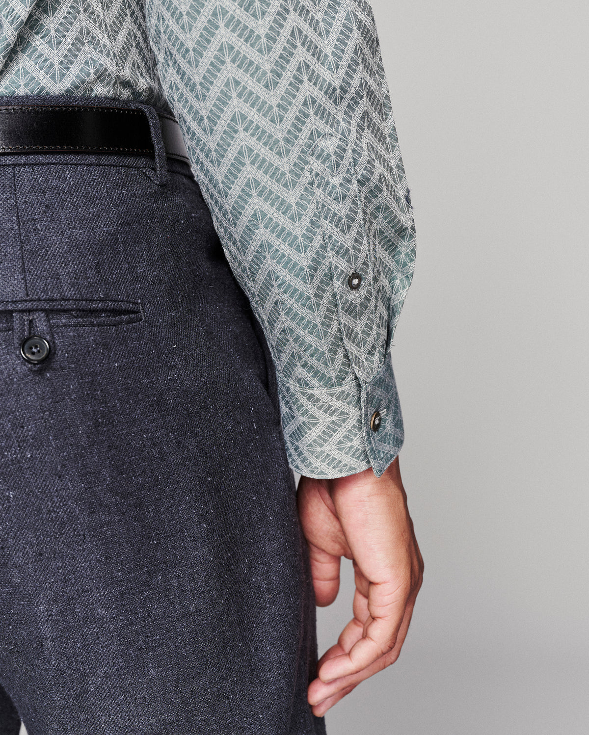 Close-up of a person wearing a patterned light blue and white long-sleeve shirt with a black belt and dark gray textured trousers, shown against a plain light gray background.