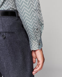 Close-up of a person wearing a patterned light blue and white long-sleeve shirt with a black belt and dark gray textured trousers, shown against a plain light gray background.