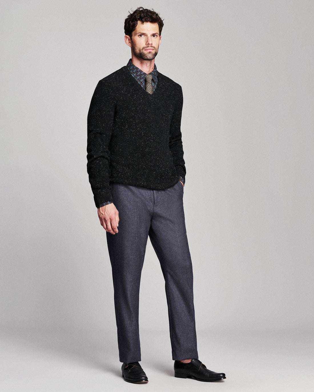 A man modeling a black knit v-neck sweater over a patterned collared shirt and tie, paired with charcoal gray dress pants and black loafers, against a light gray studio background.
