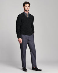 A man modeling a black knit v-neck sweater over a patterned collared shirt and tie, paired with charcoal gray dress pants and black loafers, against a light gray studio background.
