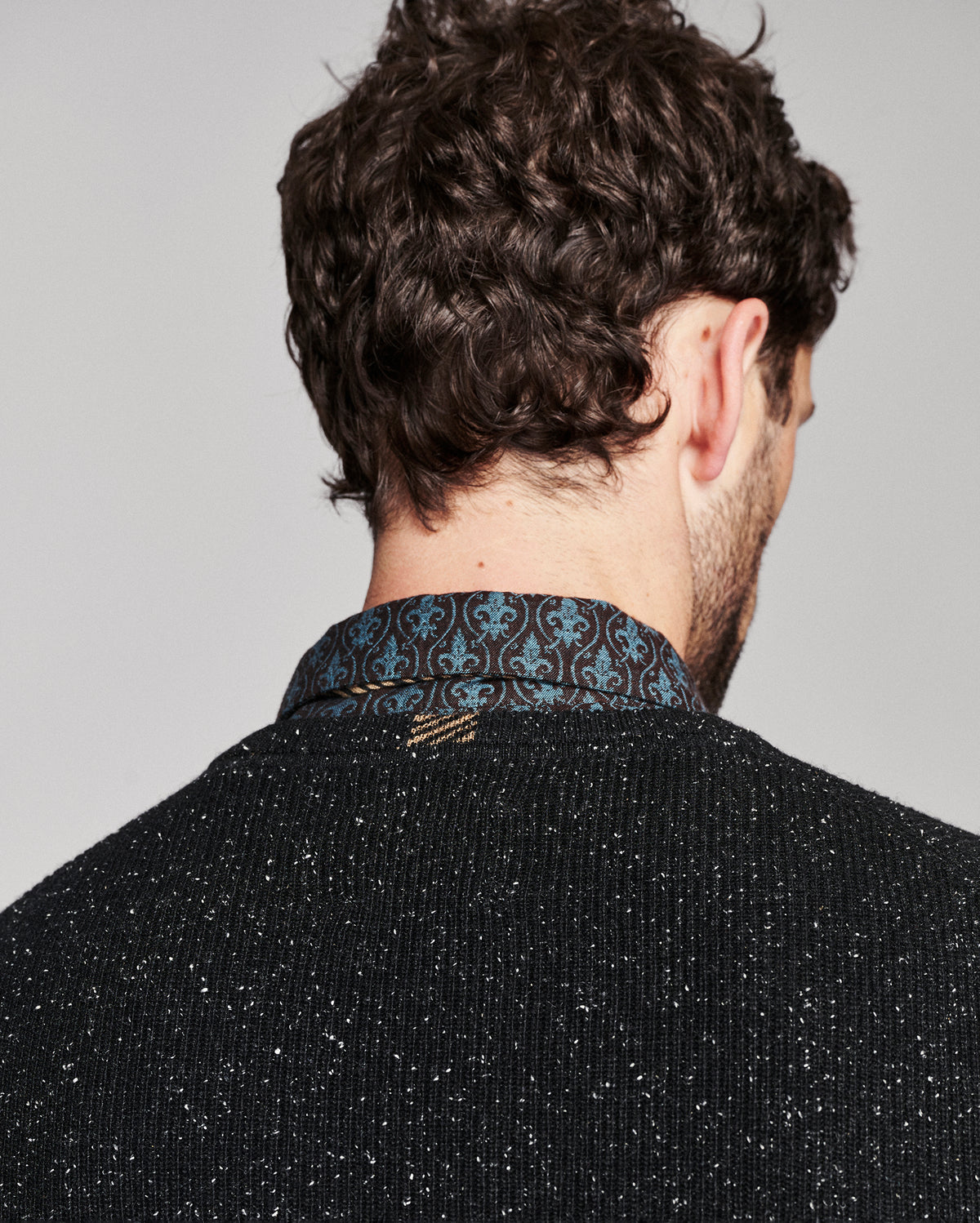 Back view of a man wearing a black speckled knit sweater and a blue and black patterned collared shirt, shown against a plain light gray background.