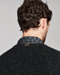 Back view of a man wearing a black speckled knit sweater and a blue and black patterned collared shirt, shown against a plain light gray background.