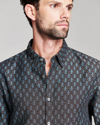 A close-up of a man wearing a dark patterned button-down shirt with a blue and brown damask-style print, photographed against a neutral gray background.