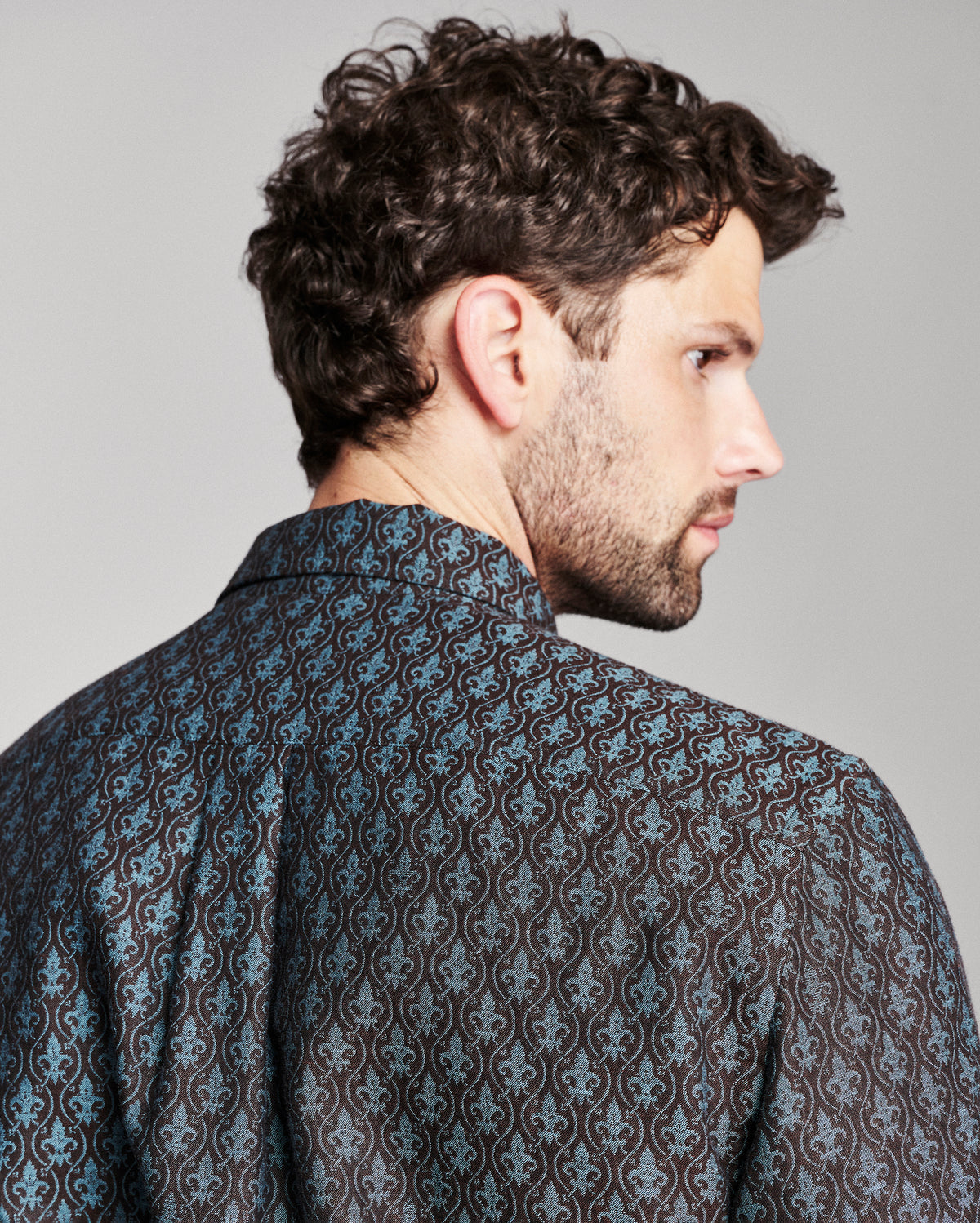 A man with curly brown hair and a beard, shown from a side-back angle wearing a dark patterned button-down shirt with blue and brown damask-style motifs, against a solid gray studio background.