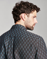 A man with curly brown hair and a beard, shown from a side-back angle wearing a dark patterned button-down shirt with blue and brown damask-style motifs, against a solid gray studio background.