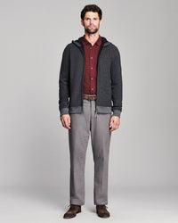 A man modeling a dark gray zip-up hoodie with a ribbed black collar, cuffs, and hem, worn over a burgundy button-down shirt and paired with gray trousers and brown shoes, against a light gray studio background.