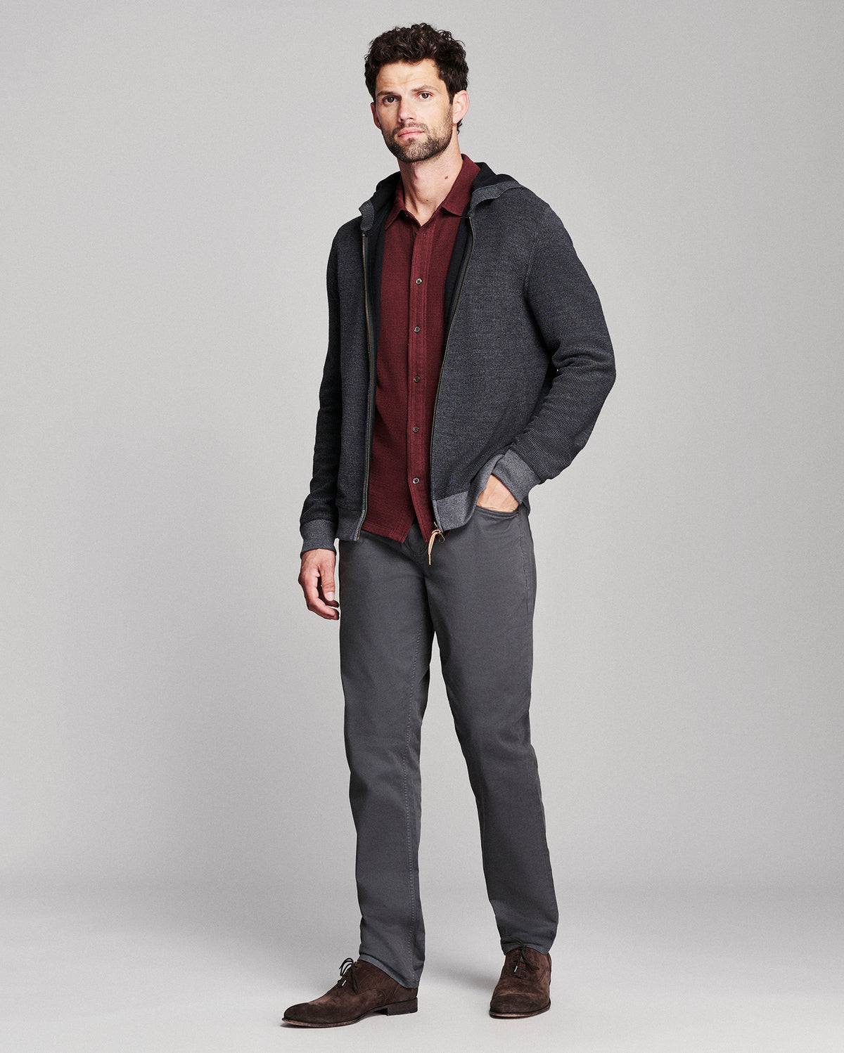 A man modeling a dark gray zip-up hoodie over a burgundy button-down shirt, paired with dark gray trousers and brown leather shoes, against a light gray studio background.