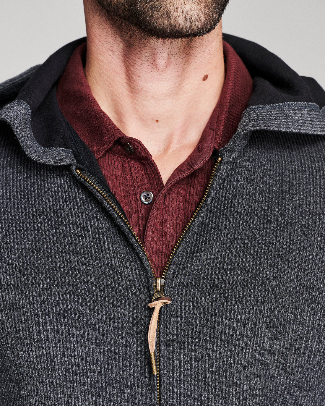 Close-up of a man wearing a dark gray ribbed zip-up hoodie with a black hood lining, layered over a burgundy ribbed button-up shirt.