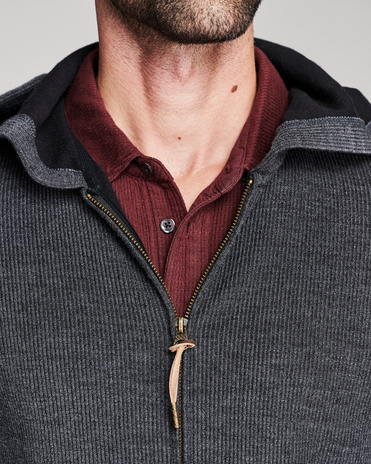 Close-up of a man wearing a dark gray ribbed zip-up hoodie with a black hood lining, layered over a burgundy ribbed button-up shirt.