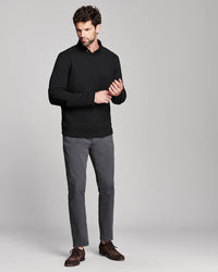 A man modeling a black long-sleeve knit polo-style sweater, paired with gray trousers and dark brown leather shoes, against a light gray studio background.
