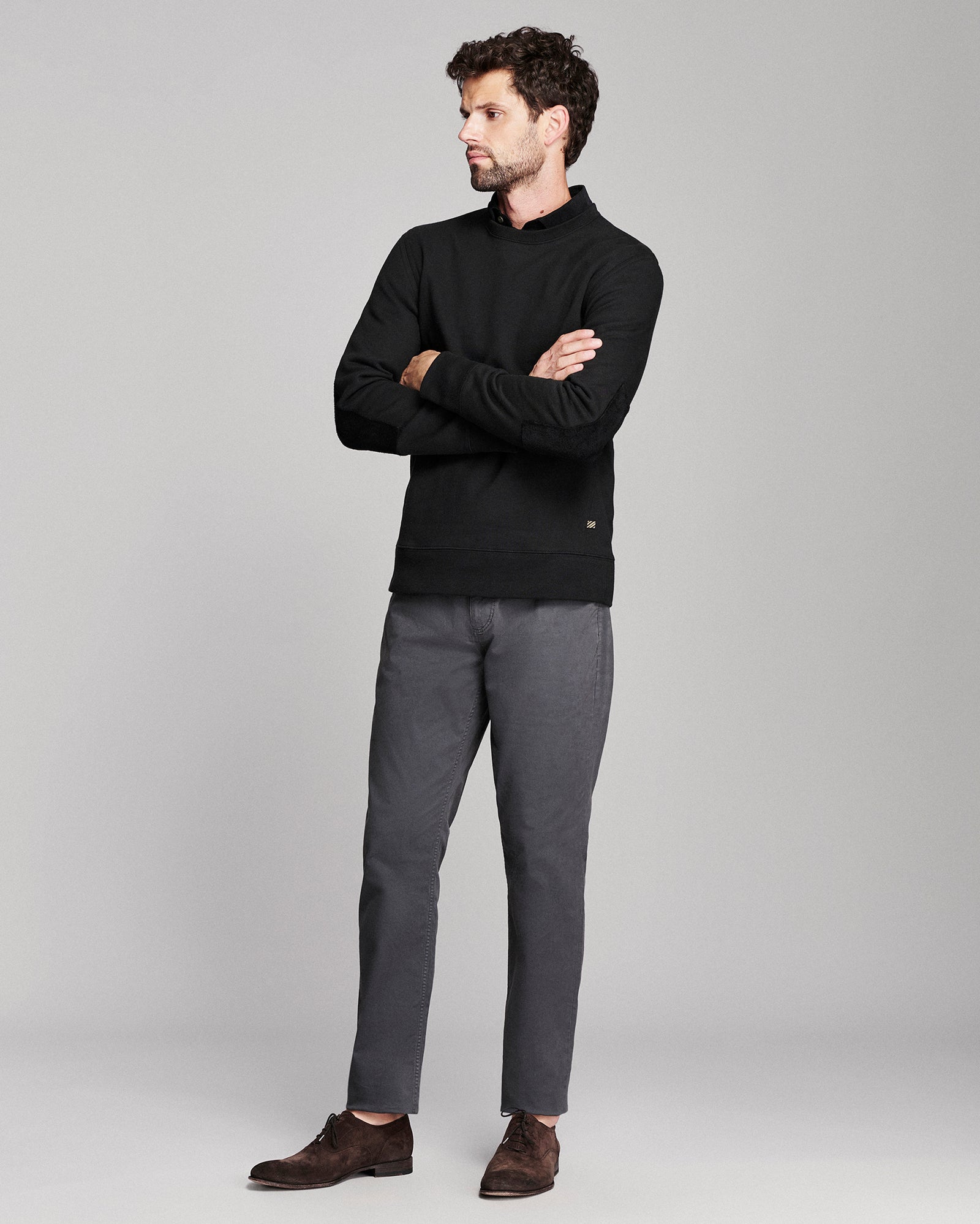 A man wearing a black long-sleeve mock-neck sweater, dark gray trousers, and brown suede loafers, standing with arms crossed against a light gray studio background.