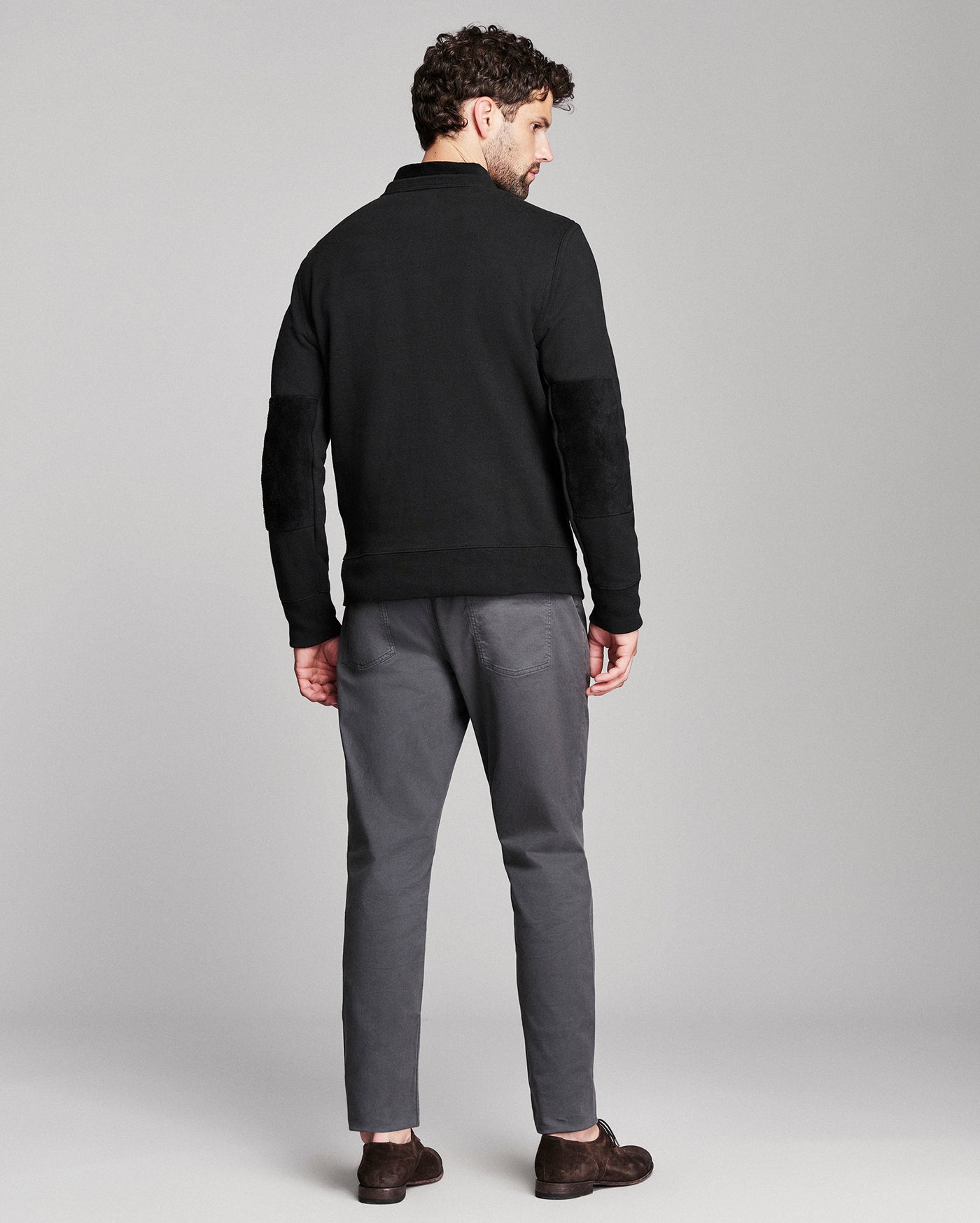 A man shown from the back wearing a black knit sweater with elbow patches, gray trousers, and dark brown shoes, photographed against a light gray studio background.