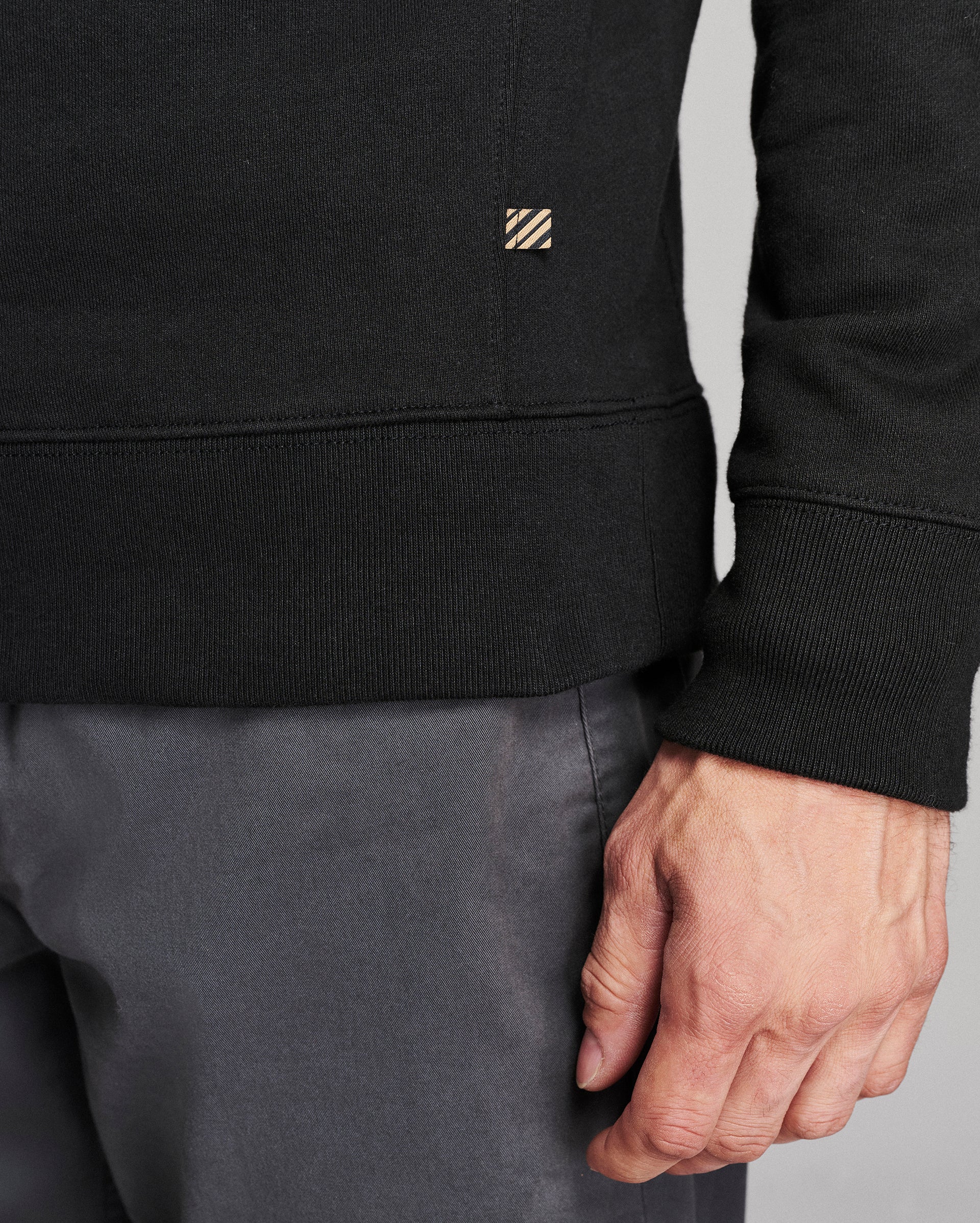 Close-up of a person wearing a black ribbed sweatshirt with a small striped square logo patch on the chest, paired with gray pants, against a light gray background.