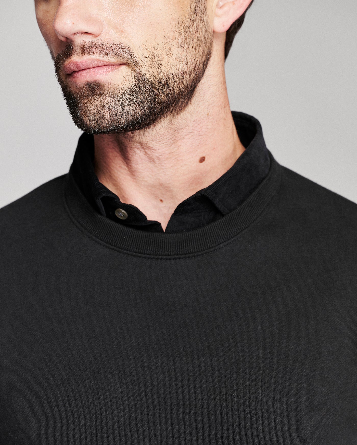 Close-up of a man wearing a black crewneck sweater layered over a black collared shirt, against a plain light gray background.