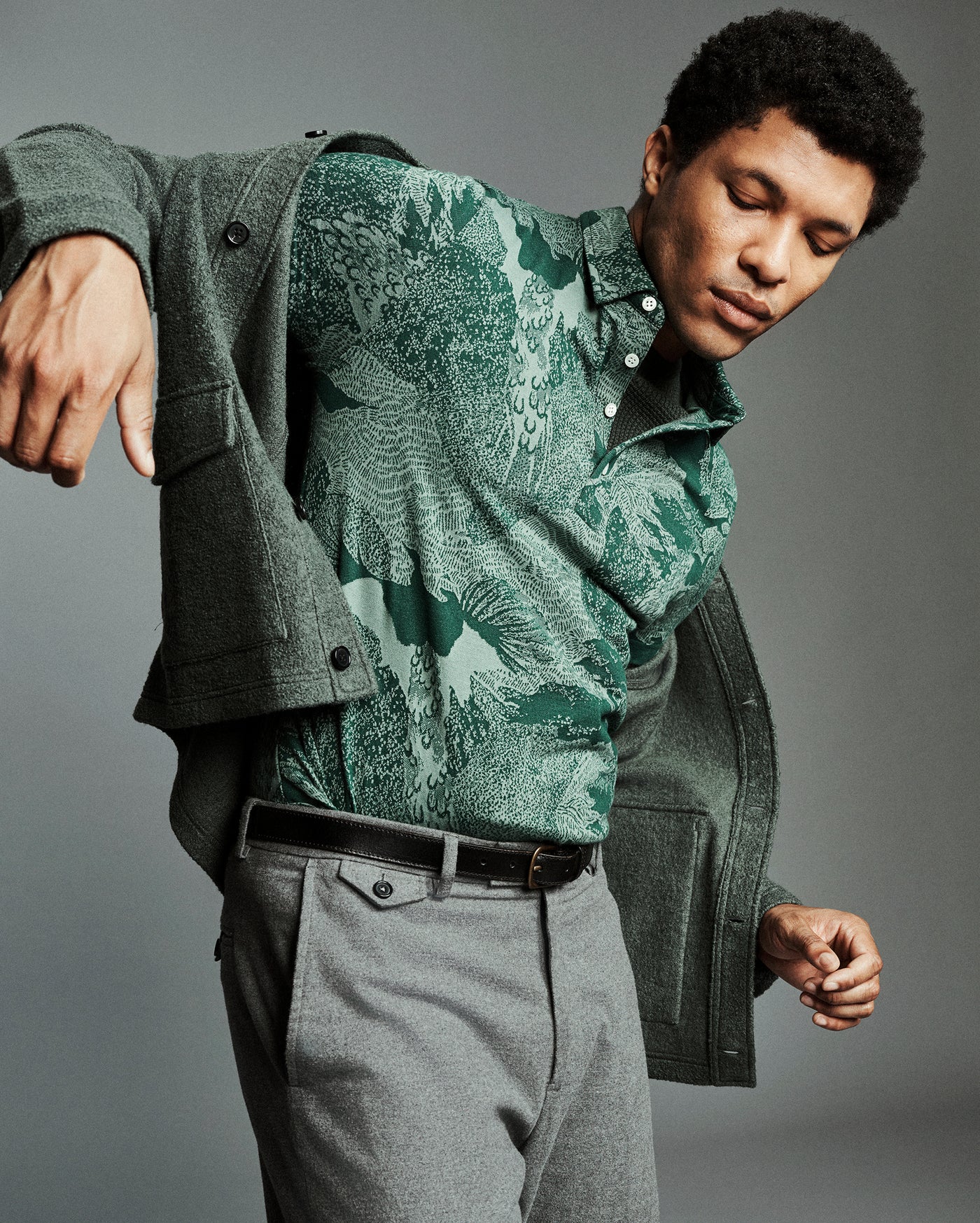 A man wearing a green patterned button-down shirt under an open dark green cardigan, paired with gray trousers and a black belt, posed against a solid gray studio background.