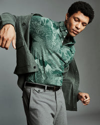A man wearing a green patterned button-down shirt under an open dark green cardigan, paired with gray trousers and a black belt, posed against a solid gray studio background.