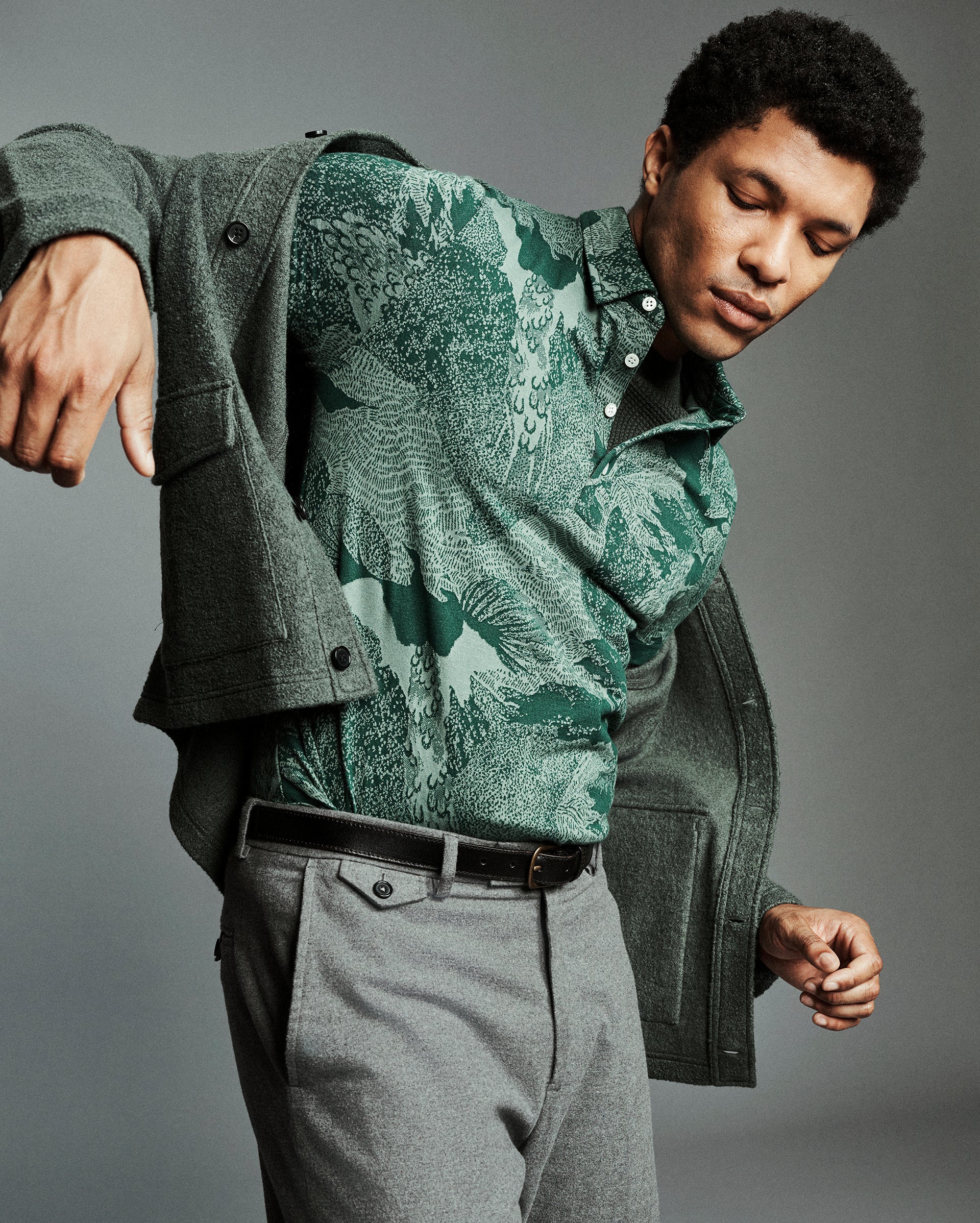 A man wearing a green patterned button-down shirt under an open dark green cardigan, paired with gray trousers and a black belt, posed against a solid gray studio background.