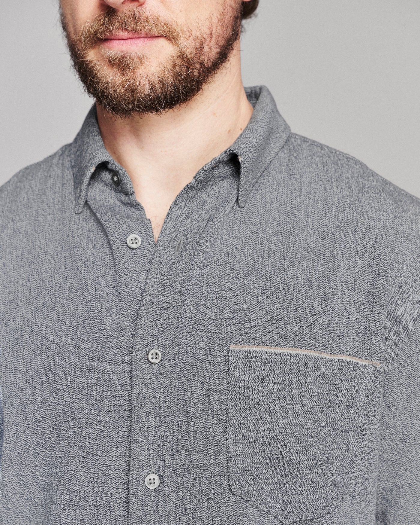 Close-up of a man wearing a textured gray button-down shirt with a chest pocket, showing the fabric weave and buttons against a neutral gray background.