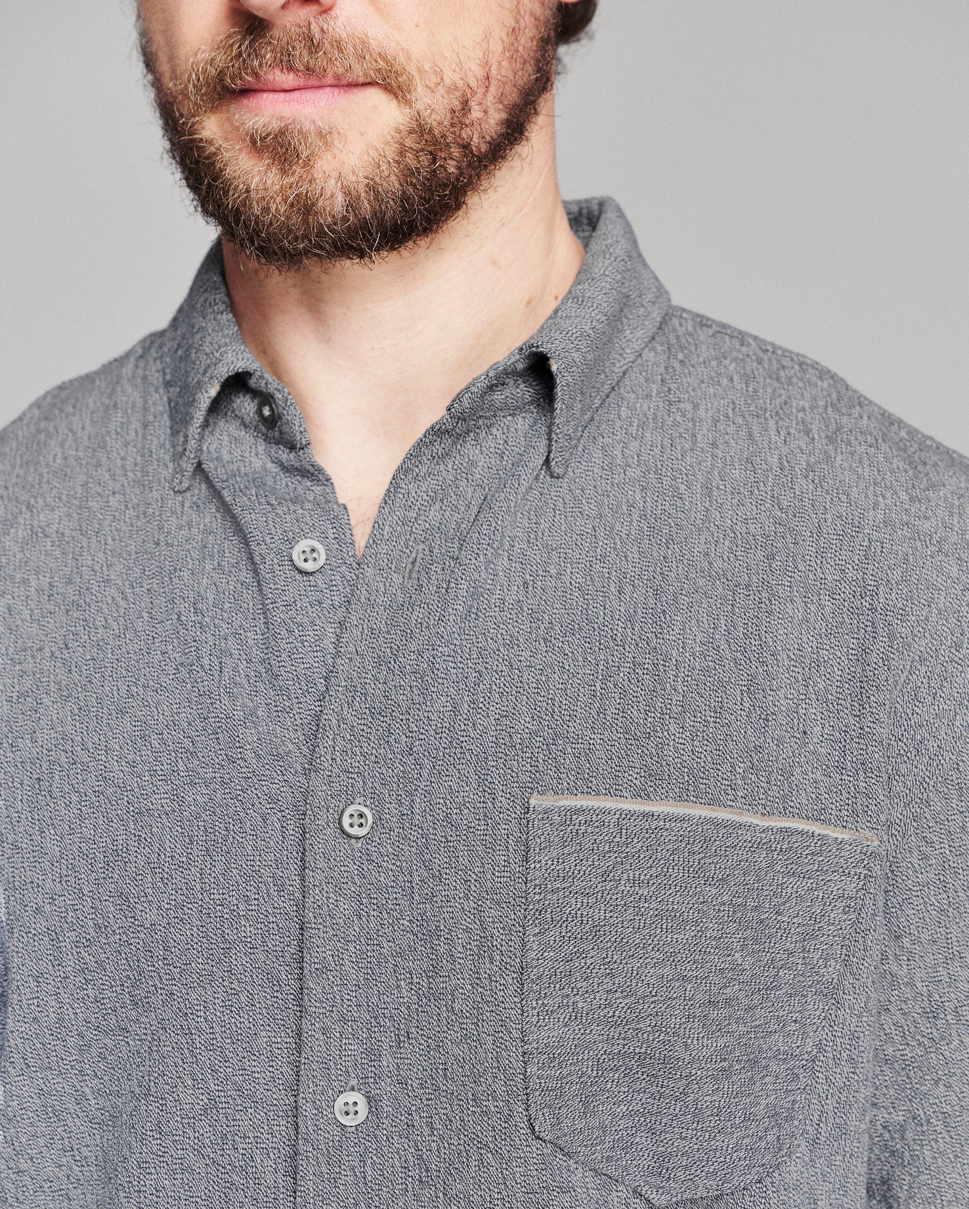 Close-up of a man wearing a textured gray button-down shirt with a chest pocket, showing the fabric weave and buttons against a neutral gray background.