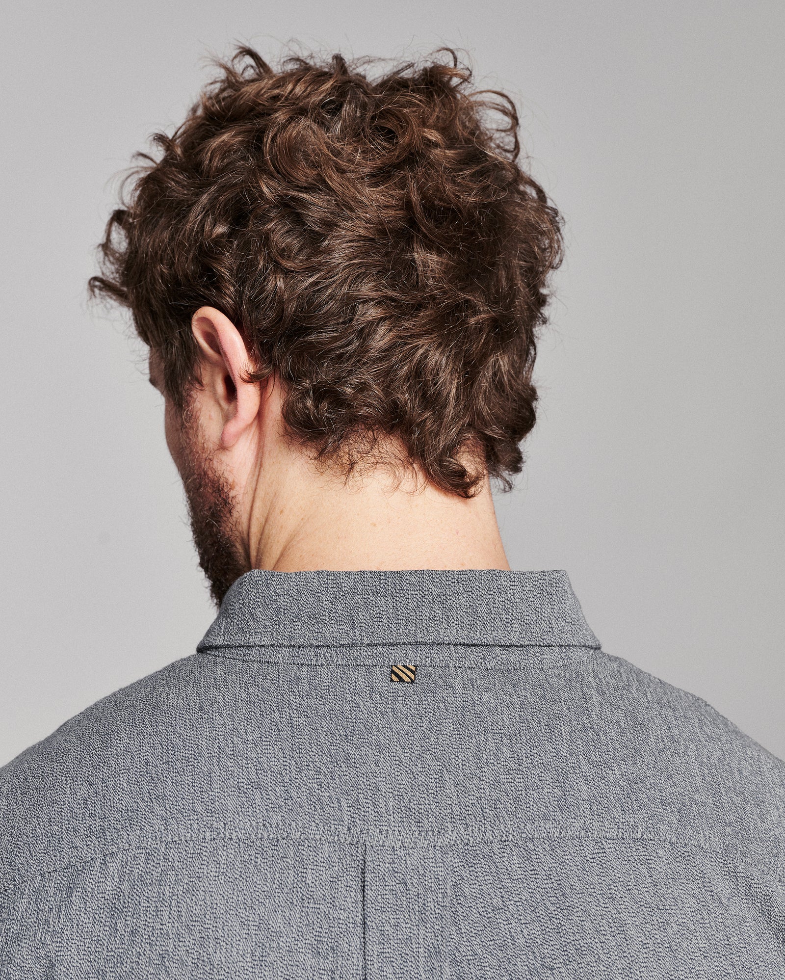 Back view of a man with curly brown hair wearing a textured gray collared shirt, showing a small gold striped button detail at the collar.
