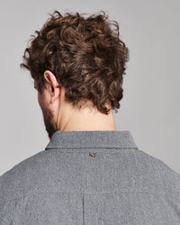 Back view of a man with curly brown hair wearing a textured gray collared shirt, showing a small gold striped button detail at the collar.