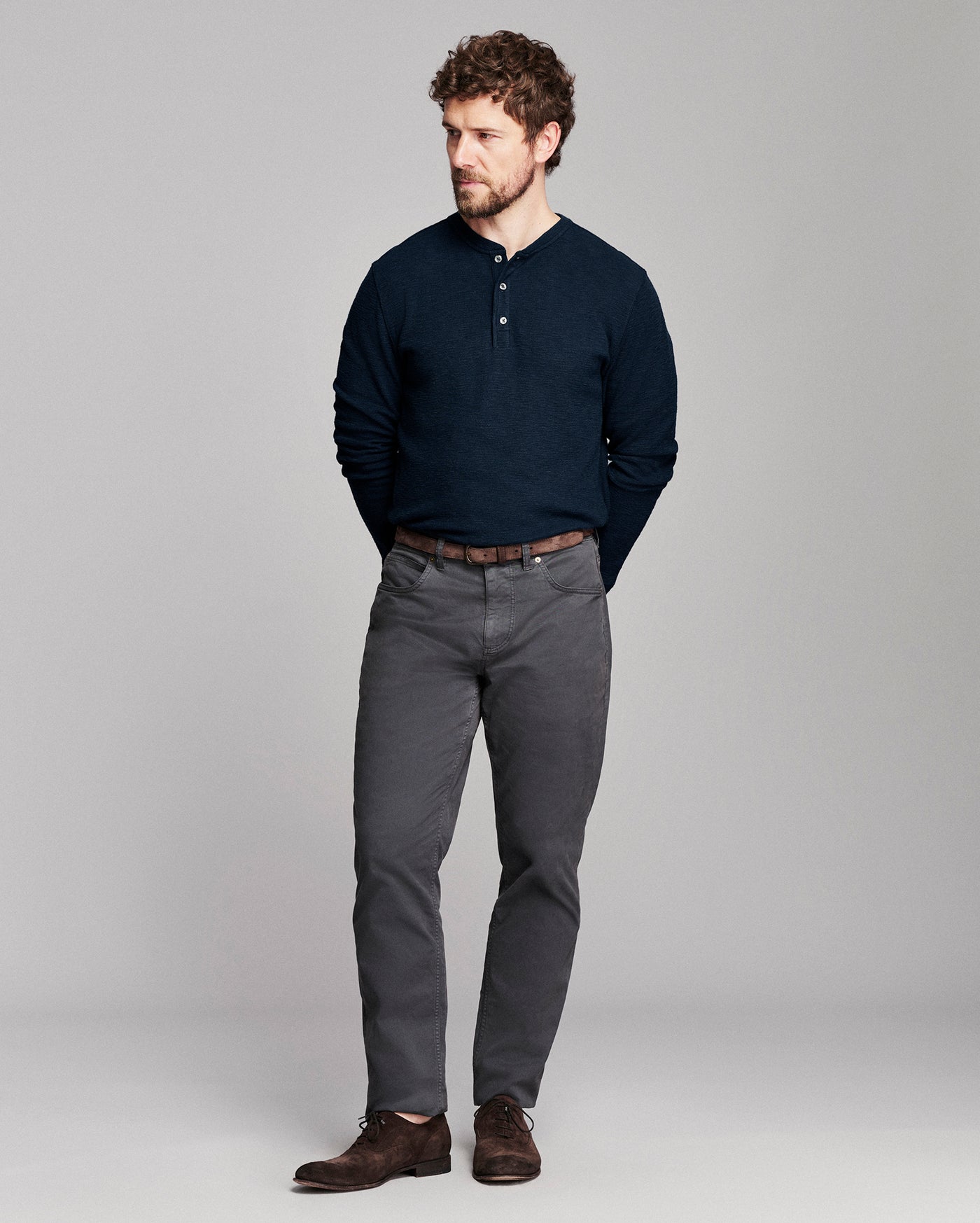 A man modeling a dark navy long-sleeve henley shirt, dark gray trousers, and brown leather shoes against a light gray studio background.
