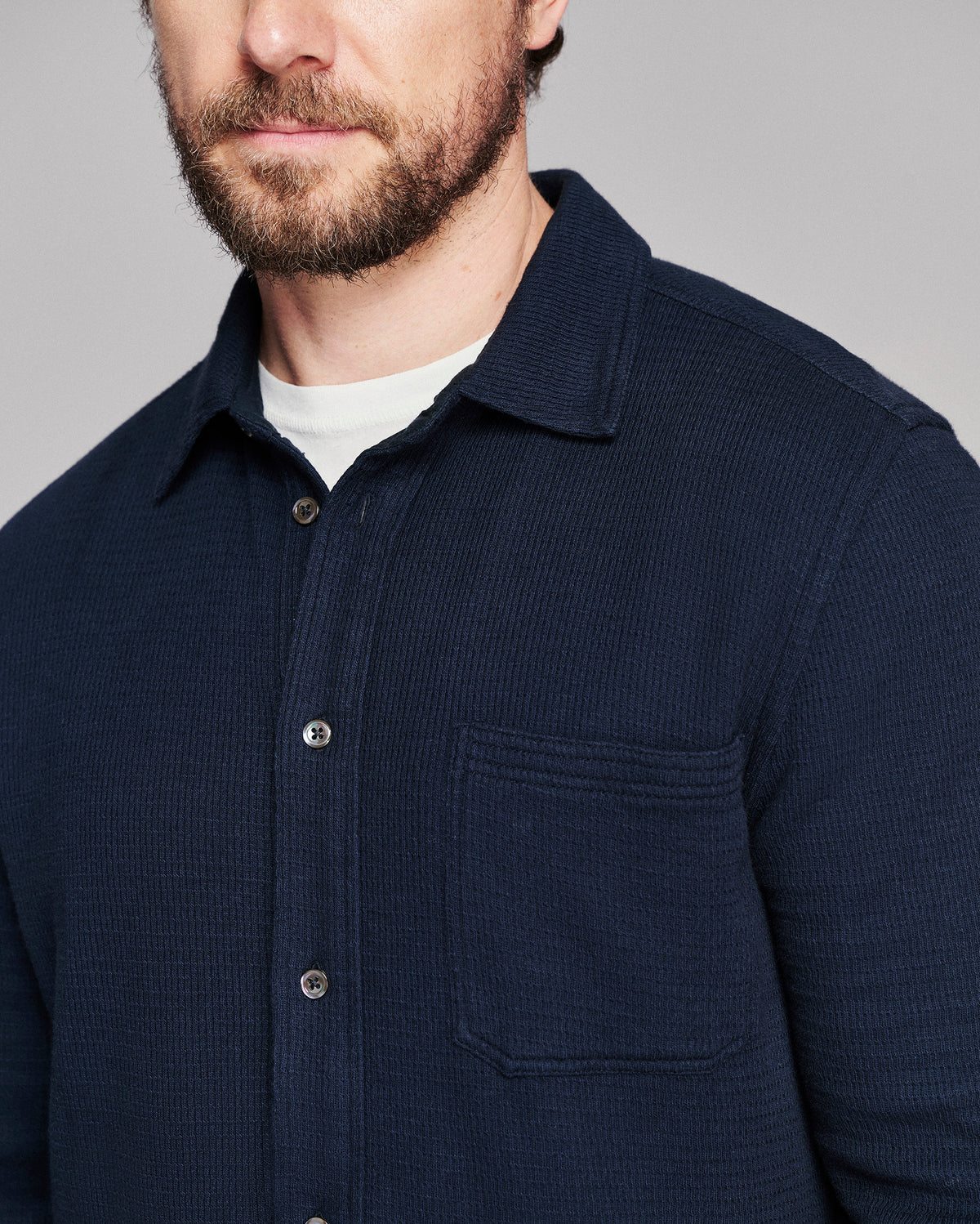 Close-up of a man wearing a dark navy textured button-up shirt over a white undershirt, against a neutral gray background.
