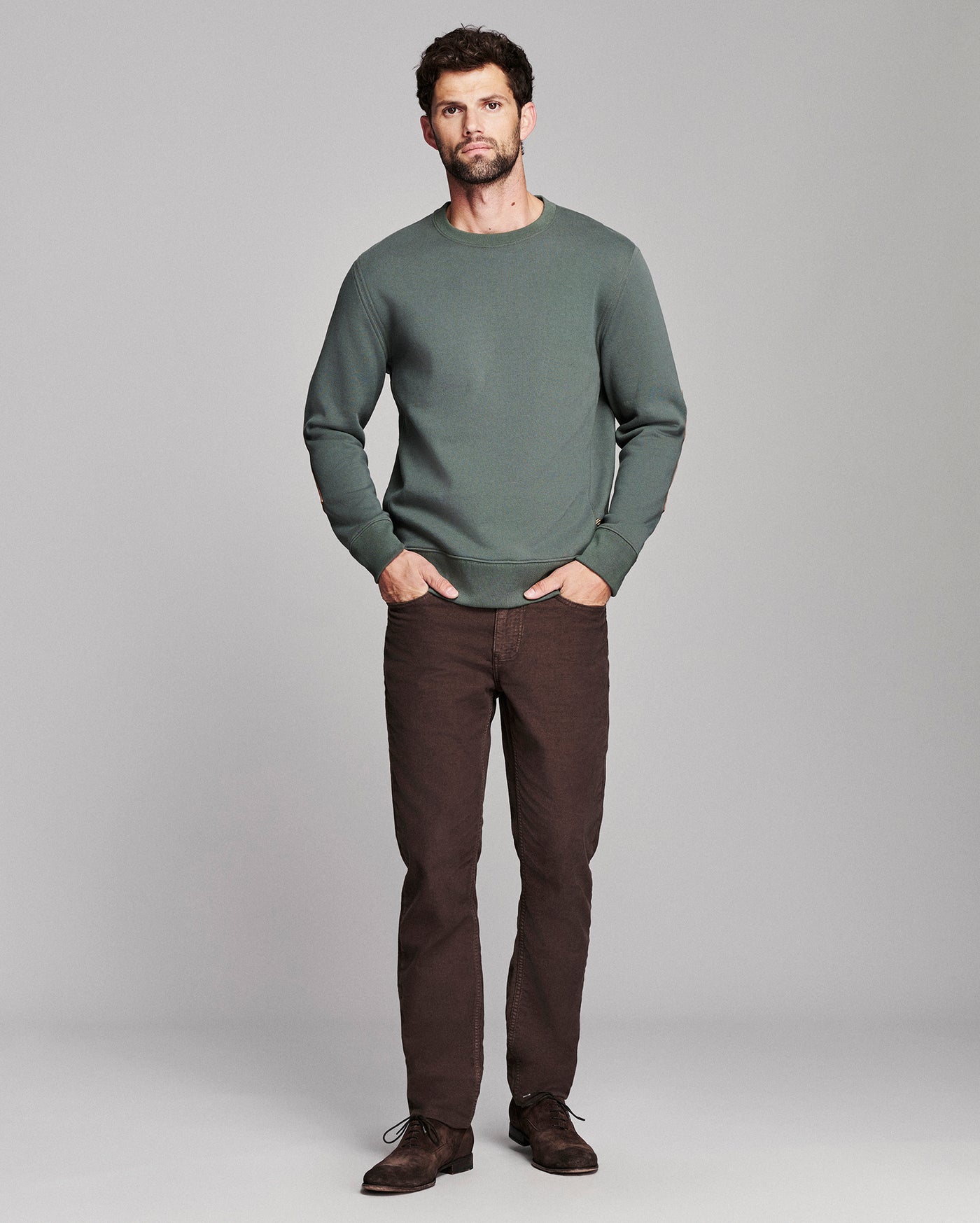 A man modeling a sage green crewneck sweater and dark brown trousers against a light gray studio background.