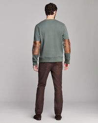 A man viewed from the back wearing a sage green crewneck sweatshirt with brown leather elbow patches and dark brown pants, set against a neutral gray studio background.