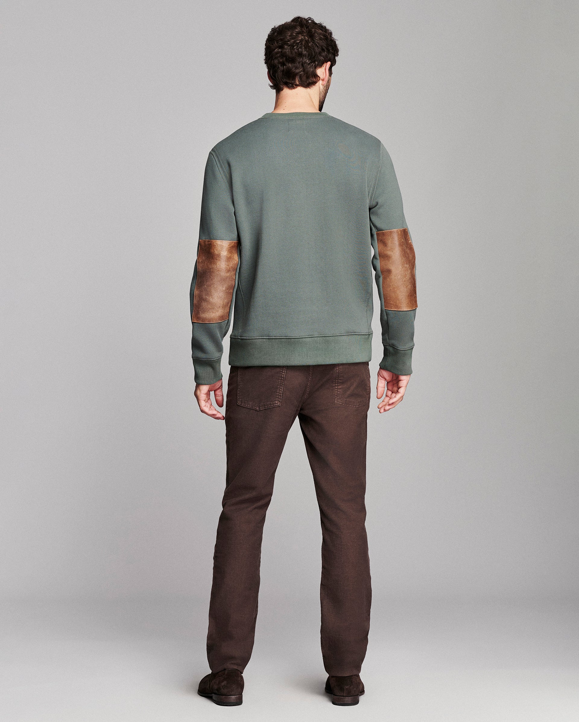 A man viewed from the back wearing a sage green crewneck sweatshirt with brown leather elbow patches and dark brown pants, set against a neutral gray studio background.