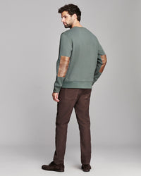 A man wearing a sage green crewneck sweatshirt with brown leather elbow patches, paired with dark brown trousers and dark brown shoes, shown from a rear three-quarter angle against a light gray studio background.