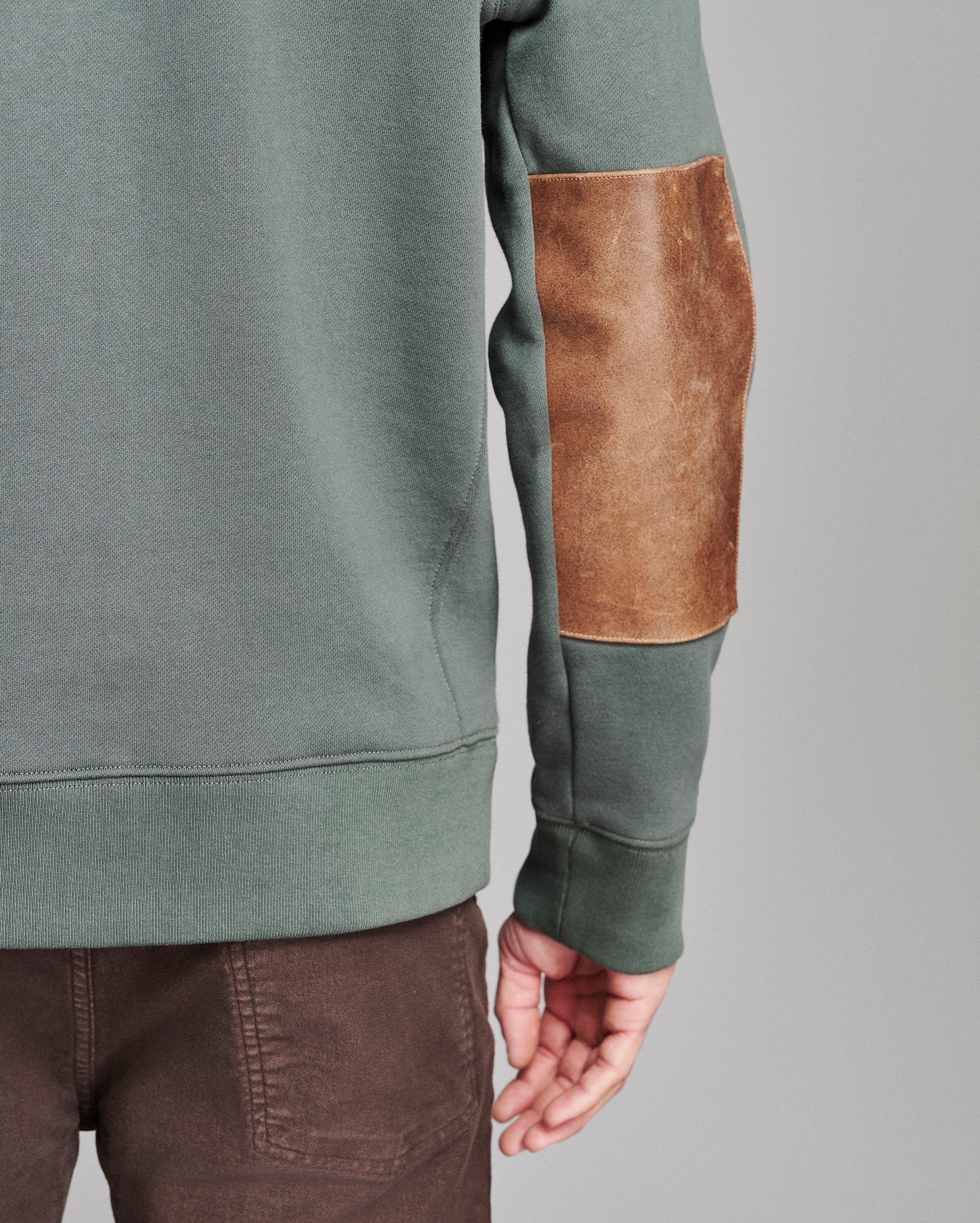 Close-up of a person wearing a muted sage green sweatshirt with a brown leather elbow patch and brown pants, shown against a plain gray studio background.